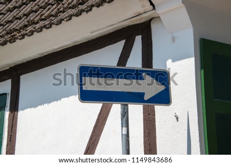 Blank white arrow on blue background street sign on pole with white wall with brown trim in background. Brown roof and green shutters
