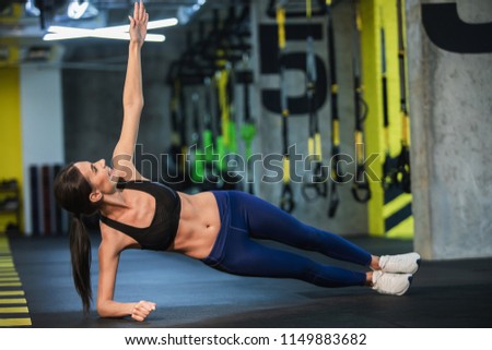 Happy lady is exercising obliques in static position in fitness studio. She is balancing on one elbow and raising other arm while looking at it. Athlete is training both strength and flexibility Royalty-Free Stock Photo #1149883682