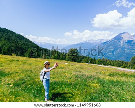 Aerial view of female tourist with backpack standing on green grass meadow and enjoy beauty of nature while photographing on smartphone. Hipster girl wanderlust recreating in summer in mountains park