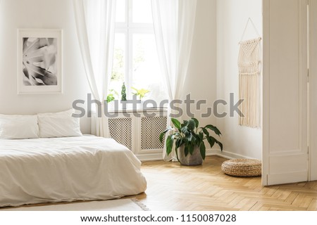 Beige macrame and a poster on the white walls of a bright, minimalist bedroom interior with herringbone hardwood floor and plants