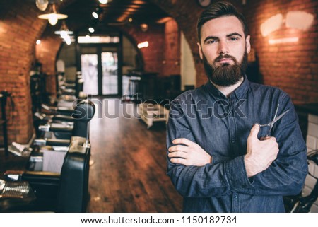 Beautiful picture of handsome guy is standing in a barbershop and keepinh his hands crossed. He is holding a pair of scissors in the right hand