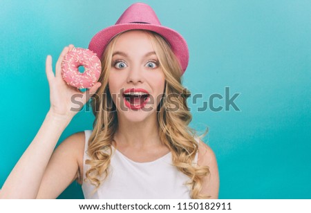 Excited girl is looking straight to camera. She is holding a big donut with pink glaze in right hand. Also young woman is keeping her mouth and eyes wide open. Isolated on blue background