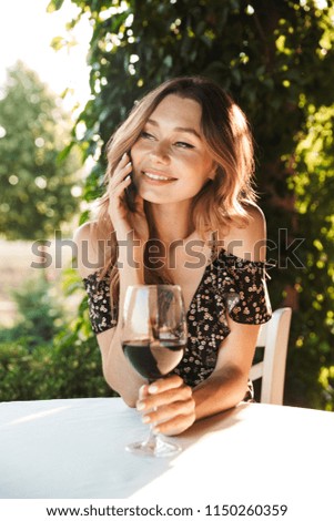 Picture of young woman sitting in cafe outdors in park holding glass drinking wine talking by mobile phone.