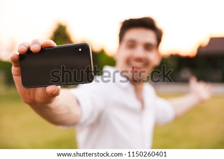 Picture of young happy man outdoors take a selfie by mobile phone. Focus on phone.