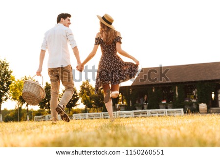 Back view picture of young loving couple with basket of drinks and food going to have picnic outdoors.