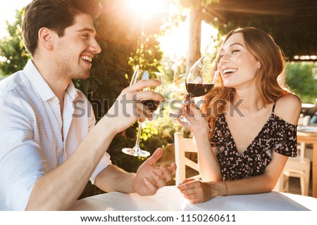 Picture of young loving couple sitting in cafe by dating outdors in park holding glasses of wine drinking talking with each other.