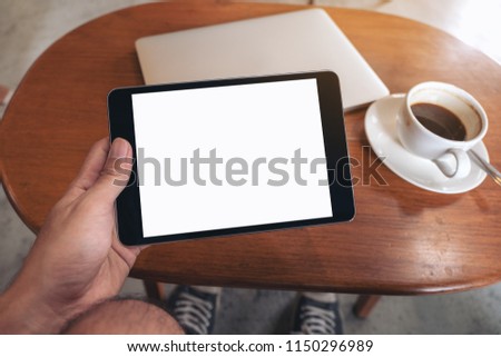 Mockup image of hand holding black tablet pc with blank desktop white screen with laptop and coffee cup on wooden table