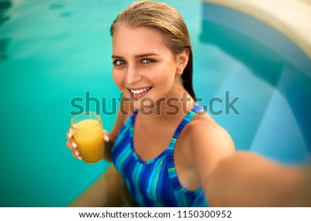 Attractive blonde woman with long hair sitting at poolside and taking selfie portrait with cellphone camera. Slim girl wear blue striped body swimsuit and holds a glass with orange juice.