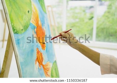 Woman hand holding brush and painting couple of swimming goldfish and lotus leaves on white canvas, water color art. Hobby idea.