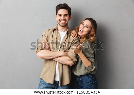 Portrait of a cheerful young couple standing together isolated over gray background, showing thumbs up