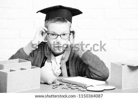 young cute pupil boy in grey sweater and glasses sitting at desk with copybook wooden numbers pink piggy pig bank in graduation cap in classroom on white brick wall background.