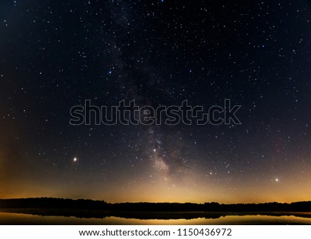Milky Way galaxy in starry night sky over lake with reflections of trees in forest with light pollution dome on horizon showing Mars, Saturn, and Jupiter. 