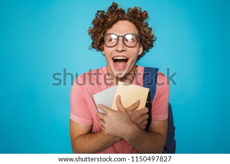 Photo of nerd student guy with curly hair wearing glasses and backpack laughing and holding books isolated over blue background