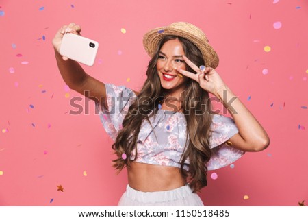 Portrait of a happy young girl in summer clothes taking a selfie under confetti rain over pink background