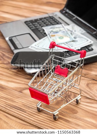 Shopping cart and laptop with money and smartphone on the table. Online shopping and Ecommerce.