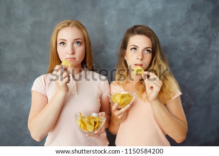 Mindless snacking, overeating, food addiction, diet, fast food. Two cute apathetic young women eating potato chips with indifferent face expression