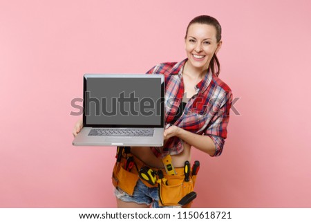 Woman with kit tools belt full of variety instruments hold laptop pc computer with blank black empty screen display touchscreen isolated on pink background. Female doing male work. Renovation concept