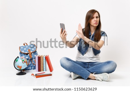 Young concerned dissatisfied woman student hold mobile phone show stop gesture with palm sit near globe backpack school books isolated on white background. Education in high school university college