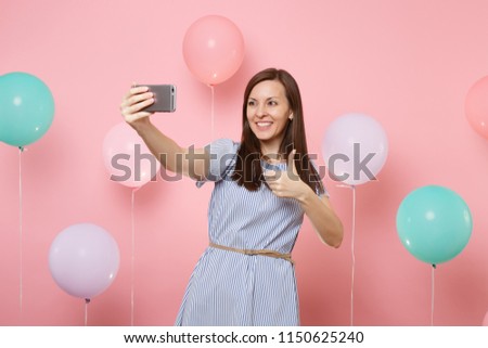 Portrait of beautiful smiling woman in blue dress doing selfie on mobile phone showing thumb up on pink background with colorful air balloons. Birthday holiday party, people sincere emotions concept