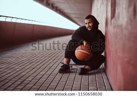 Portrait of a dark-skinned guy dressed in a black hoodie and sports shorts holds a basketball while sitting on a skateboard and leaning on wall.