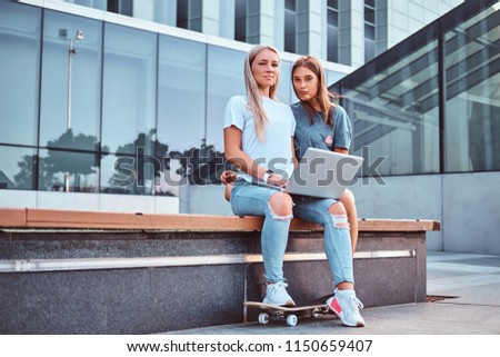 Two beautiful hipster girls sitting on the bench with a laptop on a background of the skyscraper.
