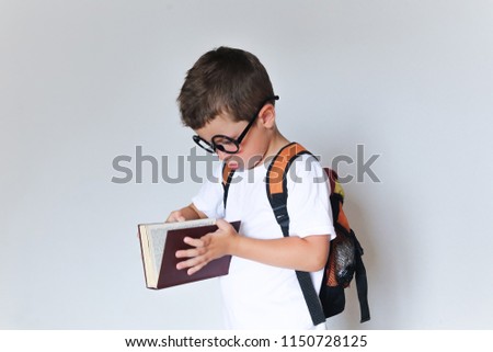 A stylish little pupil in glasses with a backpack looks into the book. The schoolboy reads the textbook at the white wall. White background. Portrait of a boy
