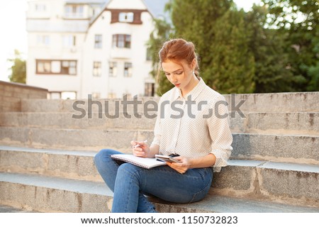 Young beautiful red hair girl with freckles sitting on a stairs near the university writing in a notebook with homework. Portrait of a student. Back to school concept