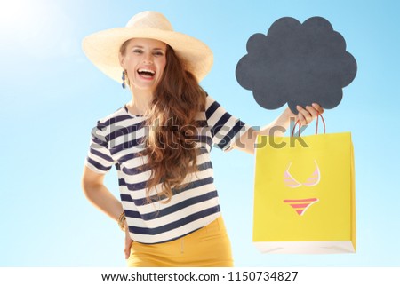 smiling young woman in straw hat against blue sky showing yellow shopping bag with bikini and blank cloud board
