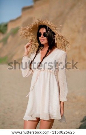 Fashion outdoor shot of gorgeous tanned young woman with dark hair in elegant summer dress and straw hat posing on summer sundy beach on background breakage in sunlight