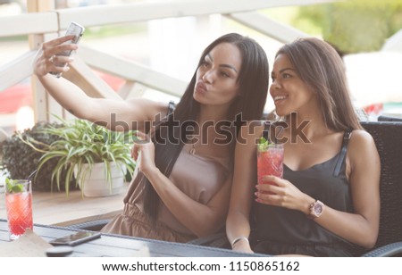 Two smiling young women sitting on a city cafe making faces while taking self portraits together with a smartphone and selfie stick