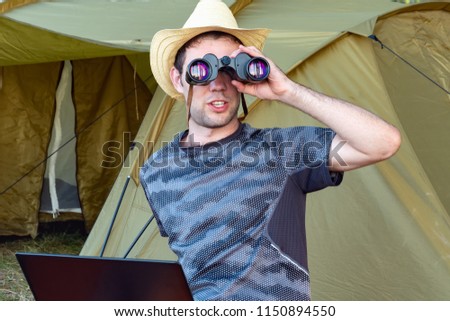 Freelance work concept. A young man in a hat sitting with a laptop near the tent and looks through binoculars. A young man working and resting in nature