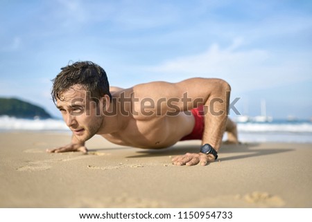 Young tanned man does a low plank on the sand beach on the sunny background of the sea with white boats and the blue sky. He wears a red swim trunks and a dark watch. Horizontal.