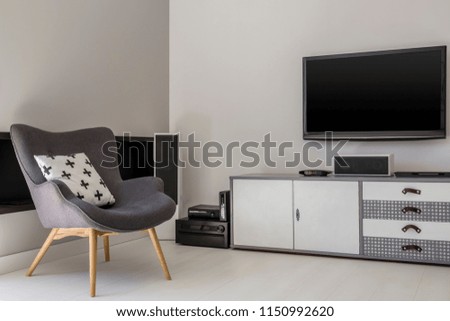 Television above cabinet next to grey armchair with patterned pillow in flat interior. Real photo