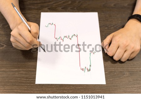 Businessman sitting at table and holds pen in his hand. There are sheet of paper with a trading chart on the table. Concept photo.