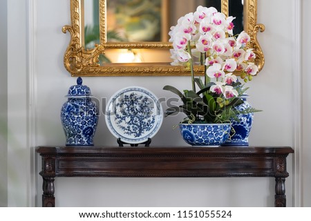 Blue paint porcelain collection with Orchid vase on a dark wooden table with golden mirror on white wall. Royalty-Free Stock Photo #1151055524