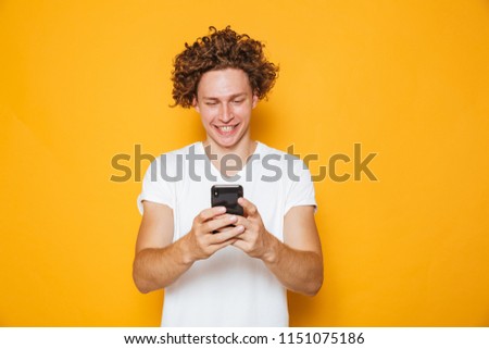 Brunette curly man in casual white t-shirt holding and using mobile phone isolated over yellow background