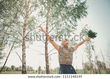 A young woman in rubber boots and a blue skirt flare with a bouquet of flowers looks up at the sky and pulls her hands up. She feels freedom, happiness and joy. Outdoors, in village on a green meadow
