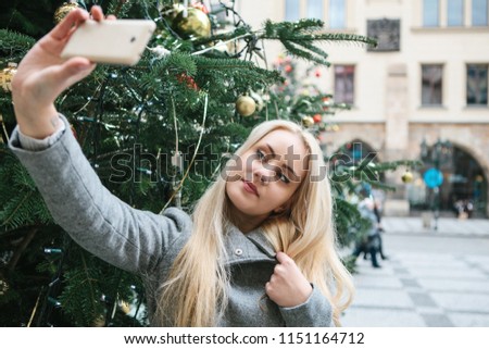 A beautiful young blonde woman or girl doing selfie or photographing next to a Christmas tree during Christmas holidays at the Old Town Square in Prague, Czech Republic.