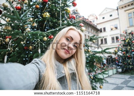 A beautiful young blonde woman or girl doing selfie next to a Christmas tree during Christmas holidays at the Old Town Square in Prague, Czech Republic.
