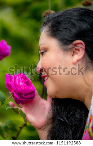 Profile portrait of a middle-aged Latin woman smelling a pink rose on a blurred green background, long black wavy hair, light makeup, enjoying a day in nature
