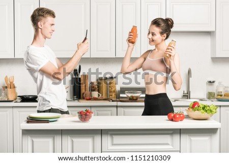 boyfriend taking photo of girlfriend with bottles of healthy juice in kitchen