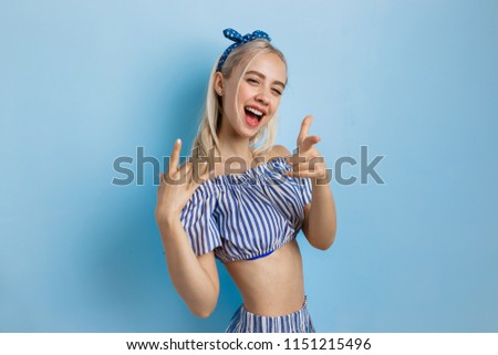 Joyful and excited beautiful blonde girl in summer outfit, pointing at camera, standing over blue background