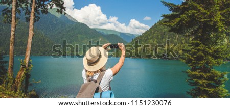 Beautiful girl traveler in a hat stands on a lake and takes pictures on a background of mountains Travel concept Summer vacation Copy space