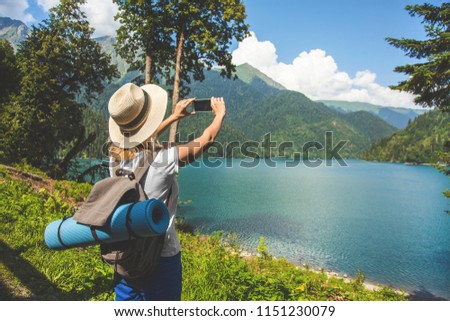 Beautiful girl traveler in a hat stands on a lake and takes pictures on a background of mountains