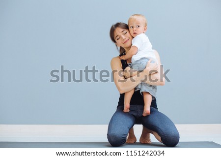 Happy sportive brunette mother hugging her lovely bay toddler son while sitting on floor over grey background. Fitness, happy maternity yoga with children concept.