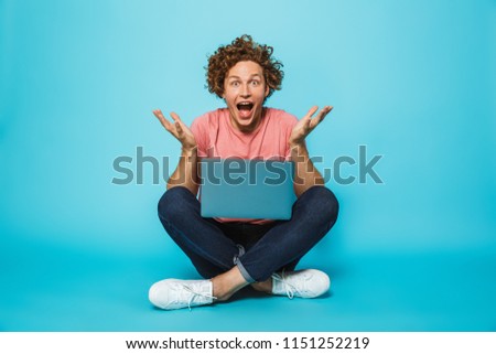 Photo of happy surprised guy 20s with brown curly hair using silver laptop, while sitting on floor with legs crossed isolated over blue background