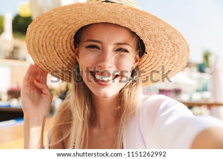 Pretty young girl in summer hat and swimwear resting at the sunny beach, taking a selfie