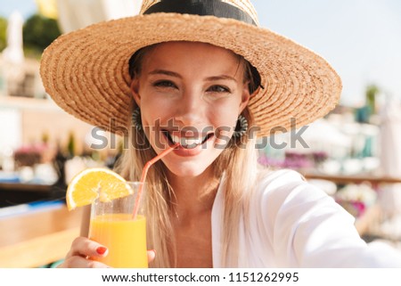 Delighted young girl in summer hat and swimwear resting at the sunny beach, sitting at a cafe with a cocktail, taking a selfie