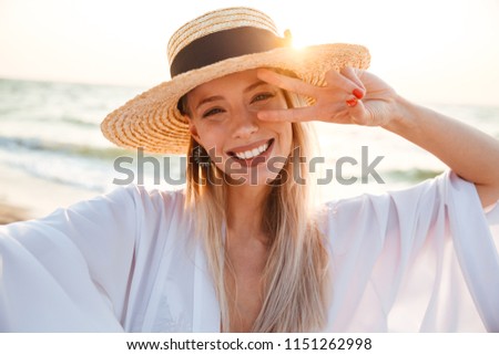 Delighted young girl in summer hat and swimwear spending time at the beach, taking a selfie, showing peace