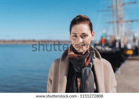 Attractive woman walking along a quay at a harbour approaching the camera with a smile with a tall ship visible behind